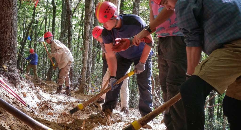 People wearing helmets use tools to work on a trail in a wooded area. 